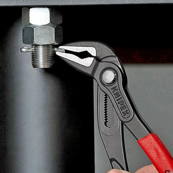 The image shows a pair of pliers gripping a metal nut that is seated on a thread. The pliers have a black body and red handles. The background is a dark surface.