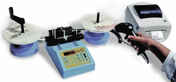 Das Bild zeigt eine Geräteanordnung zur Etikettierung. In der Mitte steht ein blaues Gerät mit Tasten, links und rechts befinden sich Rollen mit Etiketten und ein Drucker. Eine Hand hält einen Scanner.