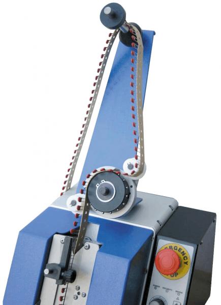 The image shows a machine with a blue casing. A movable arm with a black button and a chain leads to a large wheel. To the right, there is a red emergency stop button.