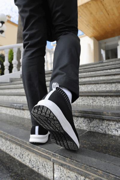 Eine Person geht die Treppe hinauf, trägt schwarze Sportchuhe mit weißen Details und schwarze Hose. Im Hintergrund sind gelbe und graue Wandflächen sichtbar.