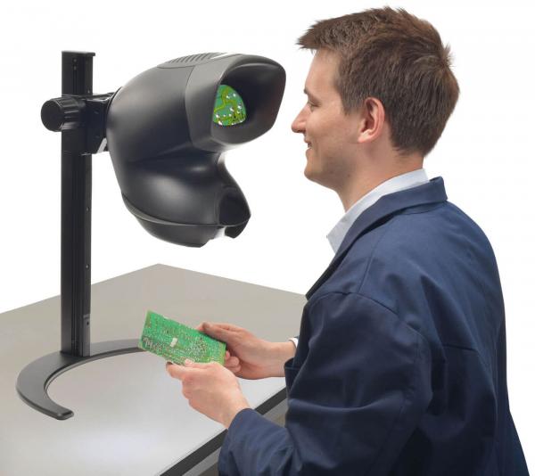 A man in a blue work coat examines a green circuit board through a large, black magnifying device. The table is plain, with the focus on the work.