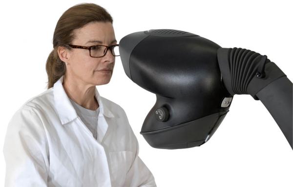 Das Bild zeigt eine Frau in einem weißen Laborkittel mit Brille, die konzentriert auf ein futuristisch aussehendes Gerät schaut. Das Gerät hat eine geschwungene Form und eine Linse.