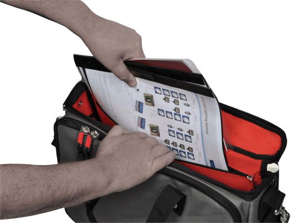 A hand reaches into a bag to pull out a document. The bag is gray with red inner pockets. The document appears to show technical information.