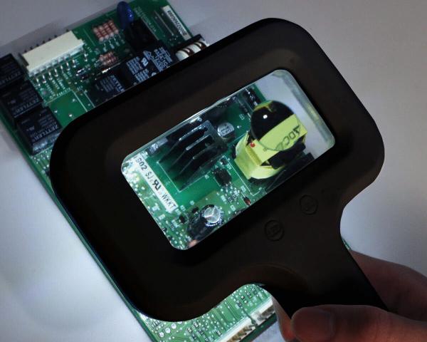 Das Bild zeigt eine Hand, die eine Lupe über eine grüne Leiterplatte hält. Die Lupe beleuchtet einen Bereich mit verschiedenen elektrischen Bauteilen, darunter ein schwarzes Element und ein gelbes.