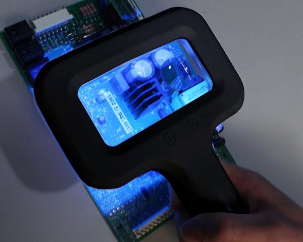 Auf dem Bild sieht man eine Hand, die ein Vergrößerungsglas mit beleuchtetem Display über eine Leiterplatte hält. Die Platine zeigt verschiedene elektronische Bauteile in bläulichem Licht.