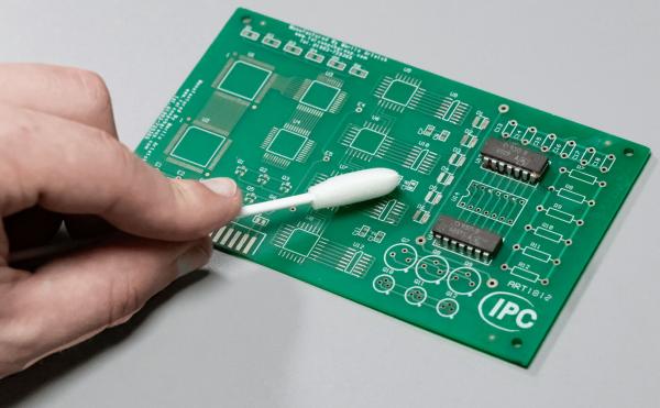 The image shows a green circuit board with various circuits. A hand is holding a cotton swab used to clean the board.