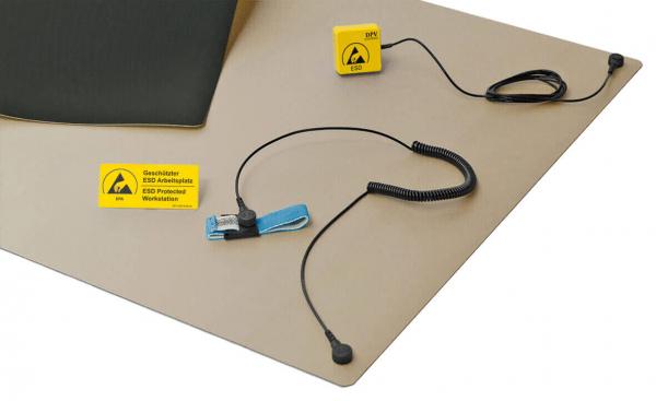 The image shows an ESD workstation mat in beige with a black cushion. Attached to it are a yellow ESD symbol and a cable with an ear clip and a connector.