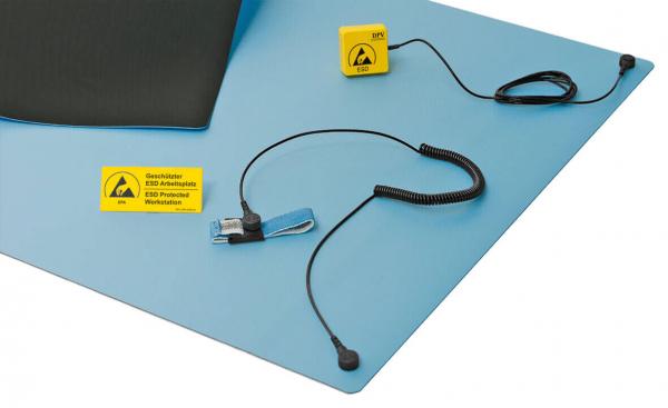 The image shows a blue ESD work mat with a black area. On it are a yellow ESD symbol, a connecting cable, and a glove with a cable for grounding protection.