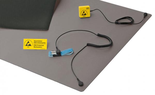The image displays an antistatic work mat in shades of gray. On the mat, there is a yellow ESD warning sign and a clamping system with cable. All parts are designed for ESD protection.