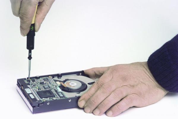 Eine Hand hält einen Schraubendreher, der in eine Festplatte gesteckt wird. Eine zweite Hand liegt auf der Festplatte, die offene Platine und einen Lüfter zeigt. Der Hintergrund ist neutral.