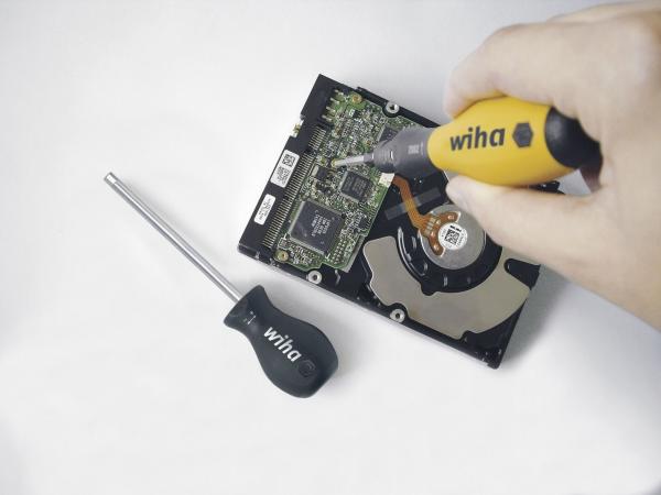 Das Bild zeigt eine Hand, die einen Schraubendreher auf eine Platine eines Festplattenspeichers richtet. Daneben liegt ein weiterer Schraubendreher mit schwarzem Griff. Der Hintergrund ist hell.