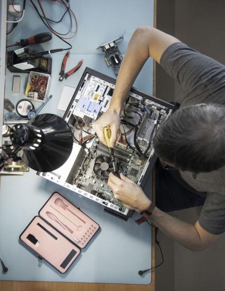 Ein Mensch arbeitet an einem offenen Computer. Mit einem Schraubenzieher wird eine Komponente montiert. Rundherum liegen Werkzeuge und Zubehör auf einem Tisch. Ein Tischlampe spendet Licht.
