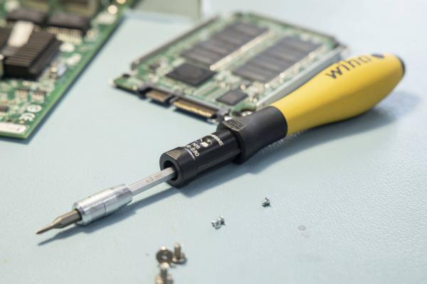 Auf dem Bild liegt ein gelber Schraubendreher mit wechselbarem Kopf auf einer Werkbank. Daneben befinden sich kleine Schrauben und eine elektronische Platine mit verschiedenen Bauteilen.