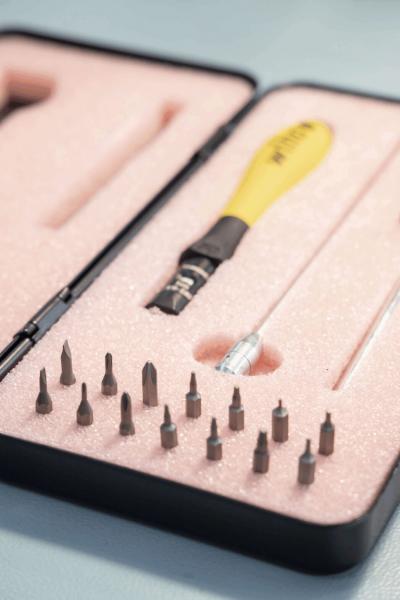 Das Bild zeigt einen Schraubenzieher mit gelbem Griff, umgeben von verschiedenen metallischen Einsätzen in einer rosa Schaumstoffunterlage. Es sind insgesamt 16 Bits vorhanden.