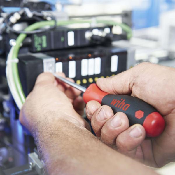 A craftsman is holding a screwdriver and working on a technical machine. In the background, cables and a control panel with small buttons are visible.