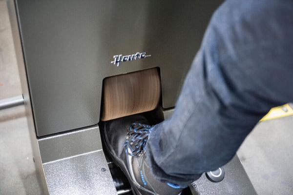 Das Bild zeigt einen Fuß, der auf ein Fußpedal drückt. Das Pedal ist an einer grauen, metallischen Maschine befestigt, die ein Logo trägt. Im Hintergrund sind unscharfe Elemente sichtbar.