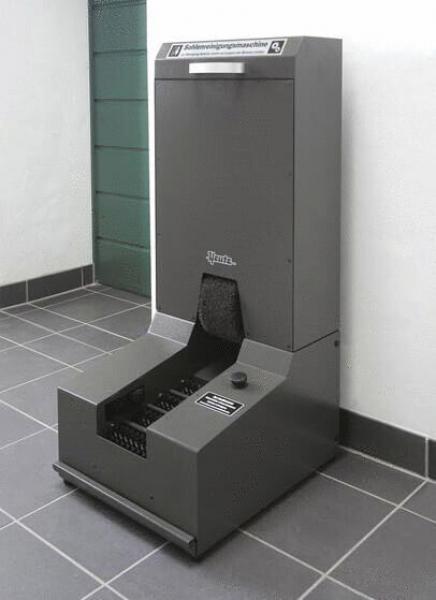 Das Bild zeigt eine graue Maschine zur Mülltrennung. Sie hat einen oberen Bereich mit einer Klappe und darunter eine breite Öffnung zur Abfallannahme, auf einem Fliesenboden.