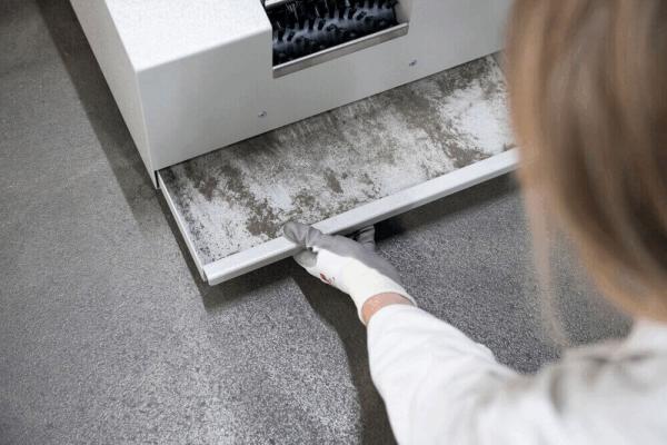 Eine Person in einem weißen Oberteil zieht eine Schublade aus einem grauen Gerät. Die Schublade ist grau und zeigt Spuren von Schmutz oder Staub. Der Boden ist ebenfalls grau.