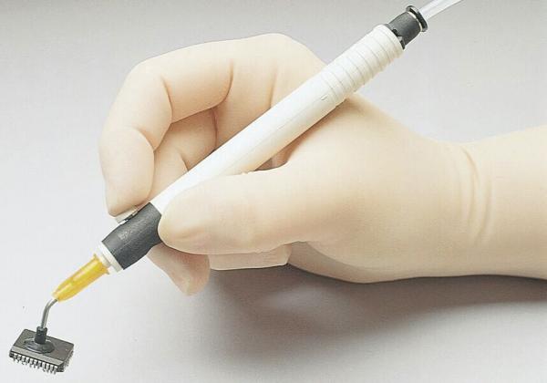 A hand in a white rubber glove holds a tool that looks like a pen. At the end of the pen is a small syringe-like opening that dispenses a clear liquid. Below it lies a black, rectangular object.