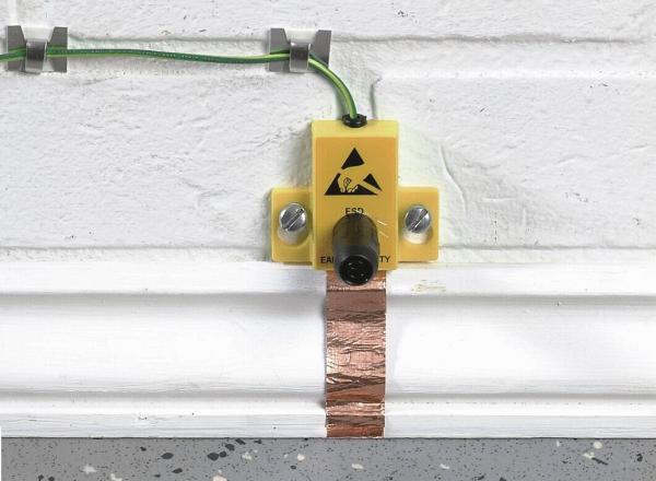 Das Bild zeigt eine gelbe Vorrichtung an einer Wand. Sie hat ein Warnsymbol und ist mit einem grünen Kabel verbunden. Darunter verläuft ein Kupferstreifen auf dem Boden.