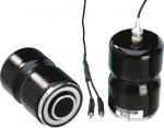 Preview: The image shows two black, round speakers with a glossy surface. They are of different heights and are connected by cables. One has a label with a serial number on it.