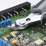 Preview: Das Bild zeigt eine elektronische Platine mit verschiedenen Bauteilen wie Widerständen und Chips. Eine Zange greift ein blauen Bauteil an der Platine.