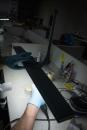 Preview: In the picture, you can see a hand with blue gloves grasping a black lamp over a bright table. In the background, various objects and desk utensils are visible.