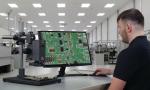 Preview: A man is sitting at a table, looking at a green circuit board with components on a monitor. The room is bright and modern, with additional workstations in the background.