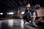 Preview: A woman is kneeling in a modern warehouse. She is wearing athletic clothing and has a determined expression on her face. Around her are wooden pallets and boxes stacked in various places.