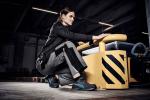 Preview: Eine sportlich gekleidete Frau kniet an einem gelben Gerät in einer Industriehalle. Sie trägt graue Arbeitskleidung und blickt konzentriert nach vorne. Das Umfeld ist modern und funktional.