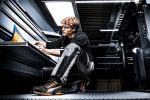 Preview: Ein junger Mann in Arbeitskleidung sitzt auf einem metallenen Boden und studiert Dokumente. Er hat lockiges, helles Haar und trägt Schuhe mit orangefarbenen Akzenten. Hinter ihm ist ein industrieller Raum.