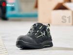 Preview: The image shows a black safety sneaker with yellow accents. It features a sturdy, reinforced toe and a quick lacing system. The shoe is on a gray floor, surrounded by packaging.