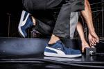 Preview: Das Bild zeigt einen Teil eines Körpers in sportlicher Kleidung, der auf einem dunklen Untergrund eine dynamische Pose einnimmt. Die Schuhe sind dunkelblau mit weißen Akzenten.
