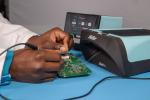 Preview: A hand works on a green circuit board, surrounded by technical devices. A device in light blue stands next to it, used for maintenance or testing.