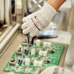 Preview: A hand in a white glove holds a pair of pliers and works on a green circuit board with various components on it. In the background, an industrial environment is visible.