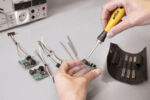 Preview: A hand holds a screwdriver over an electronic circuit board. Next to it, several cables and more circuit boards are lying around. In the background, a measuring device and a tool holder can be seen.
