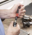 Preview: A hand holds a screwdriver with a red handle. The tool is used for working on metal parts that are lying on a table. The background is blurry.