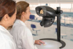 Preview: Two women in lab coats are standing in front of a special device that illuminates an object. The device has a round shape and displays enlarged details of a small object lying on a table.