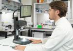 Preview: A man in a lab coat is sitting at a table, looking at a monitor connected to a microscope. In the background, shelves with laboratory utensils are visible.