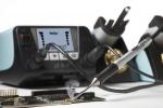Preview: The image shows a soldering iron tool being used on a circuit board as smoke rises. In the background, there is a soldering station with digital displays.