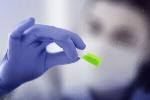 Preview: A hand with blue gloves is holding a small, brightly green object. In the background, a person with a face mask can be seen out of focus.