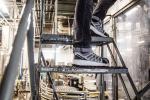 Preview: Ein Paar sportliche Schuhe in Schwarz und Grau steht auf einer Metalltreppe in einem industriellen Raum mit Maschinen im Hintergrund, der hell erleuchtet ist.