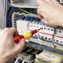 Preview: A person is working on a control cabinet with electrical components. In one hand, they are holding a screwdriver. Several fuses and cables are visible. A technical environment.