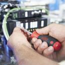 Preview: A craftsman is holding a screwdriver and working on a technical machine. In the background, cables and a control panel with small buttons are visible.