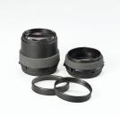 Preview: The image shows two black camera lenses with gray rubber coating. Next to them, there are two black filter rings. The objects are arranged on a light surface.