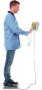 Preview: A man in a blue coat is standing on a scale. With one hand, he touches a device that is connected by a cable. The man is wearing jeans and black shoes.